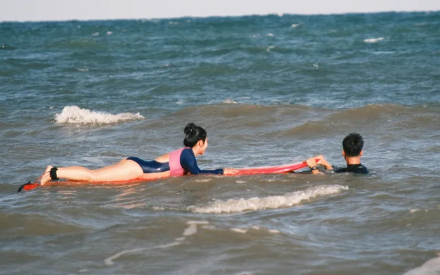 A coach guides a student practising paddling on a red surfboard in the water.