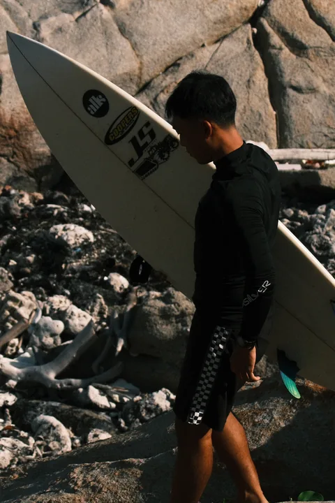A surfer carries a board along a rocky shore during a surf trip.
