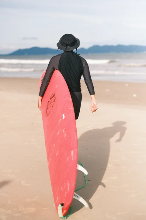 A surfer carries a red board down the beach toward the water.