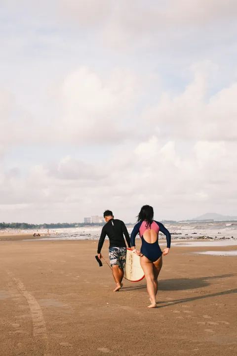 A coach and student walk back along the beach carrying a surfboard.